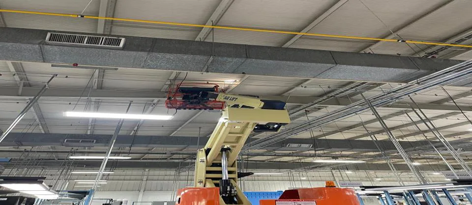 Brazo articulado elevado hacia un ducto rectangular bajo el techo de una nave