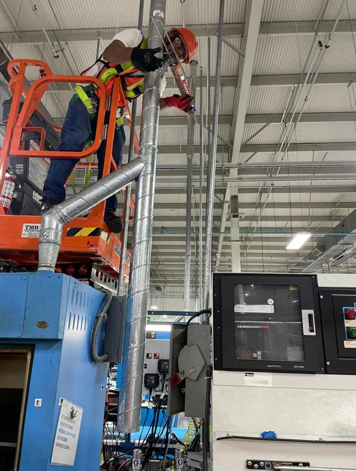 Técnico con casco naranja y arnés instalando espiroducto sobre una plataforma junto a maquinaria industrial